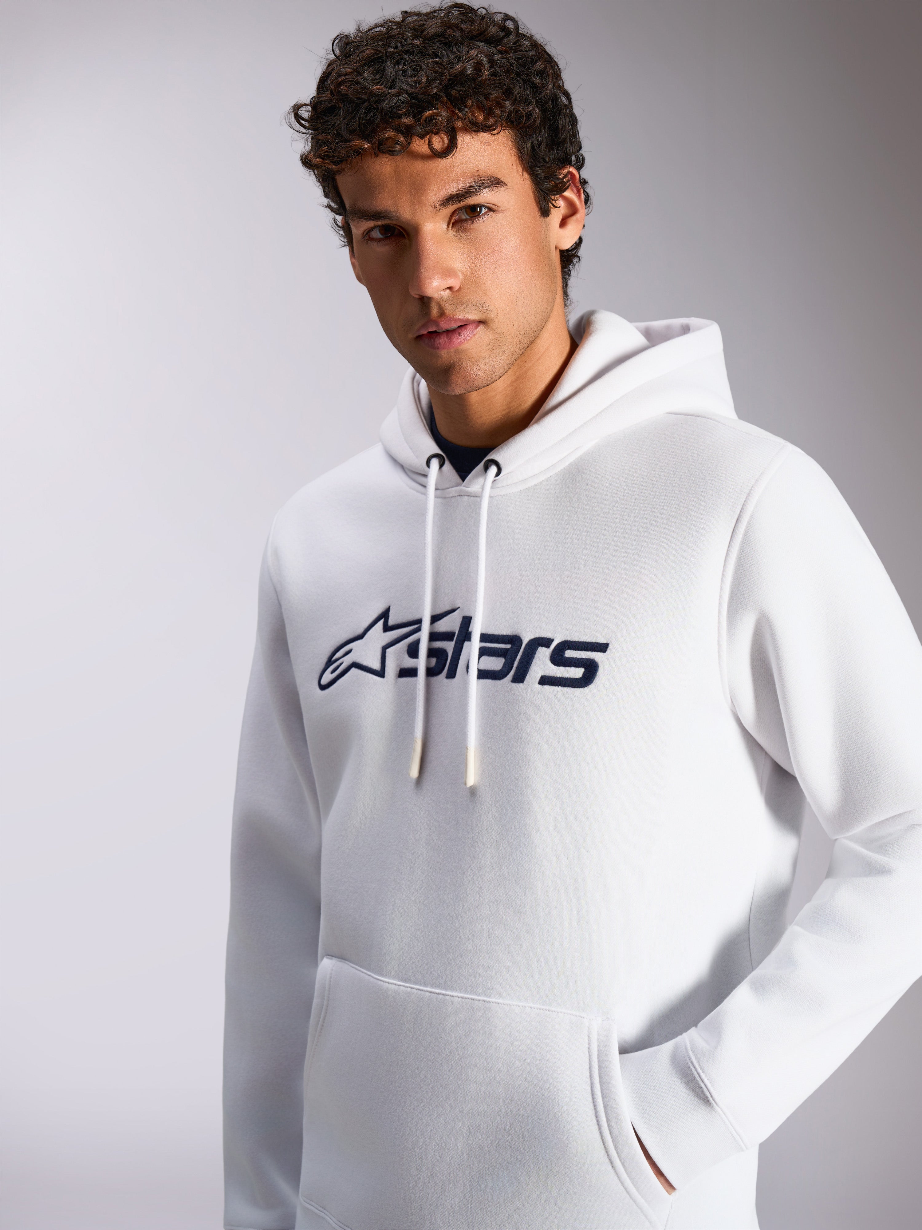 Alpinestars Blaze V3 Hoodie, camisola com capuz branca e azul marinho, usada por um modelo masculino, vestuário desportivo casual, logótipo frontal Alpinestars em azul marinho, capuz com cordão, bolso canguru, design moderno de estilo de vida