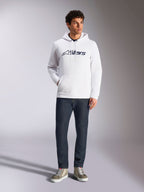 Alpinestars Blaze V3 Hoodie, camisola com capuz branca de estilo streetwear casual, com o logótipo Alpinestars azul-marinho no peito, usada por um modelo com calças de ganga azul-escuras e sapatilhas beges, com capuz ajustável e bolso frontal tipo canguru