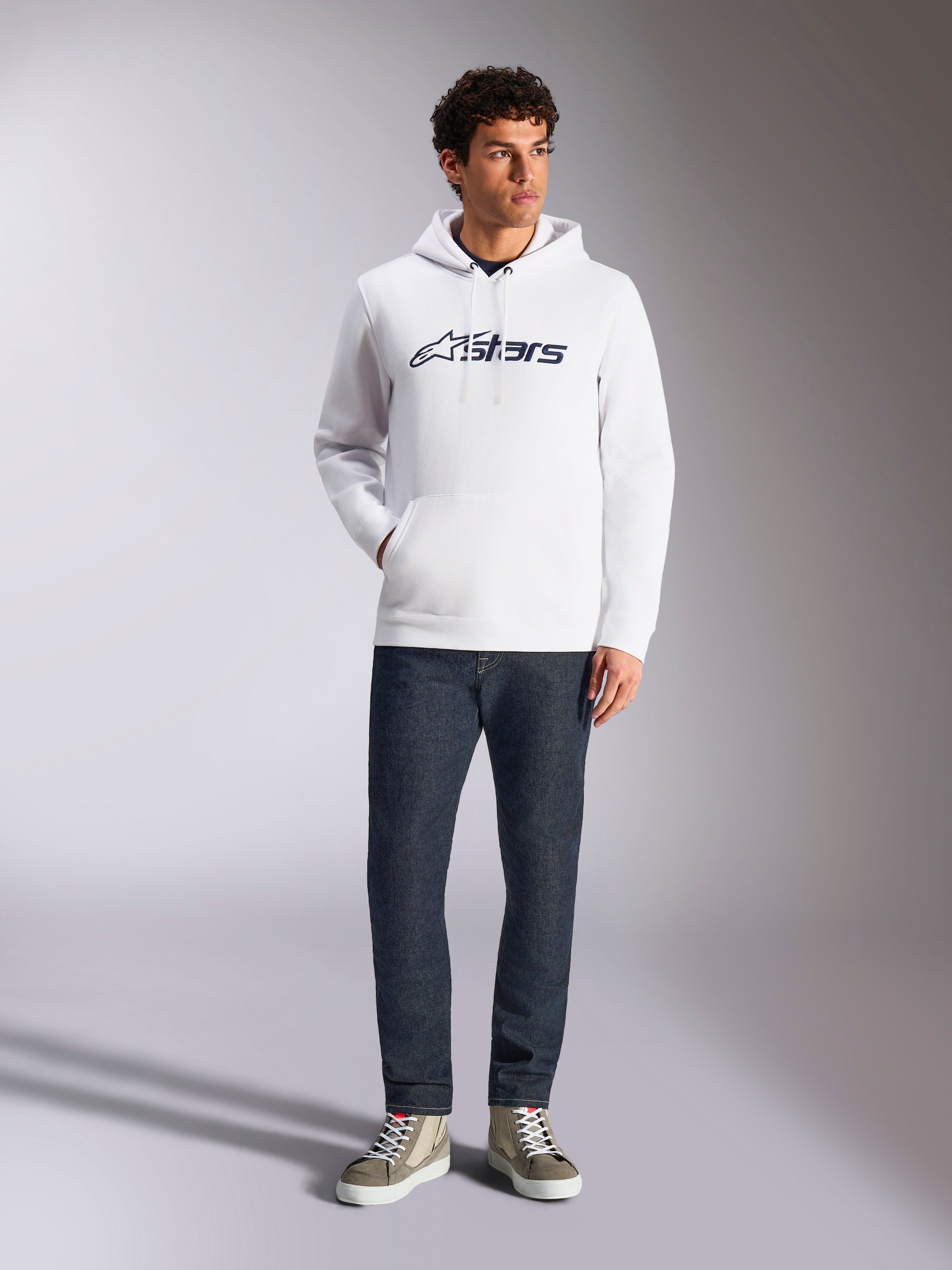 Alpinestars Blaze V3 Hoodie, camisola com capuz branca de estilo streetwear casual, com o logótipo Alpinestars azul-marinho no peito, usada por um modelo com calças de ganga azul-escuras e sapatilhas beges, com capuz ajustável e bolso frontal tipo canguru
