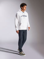 Alpinestars Blaze V3 Hoodie, branco e azul-marinho, streetwear casual usado por um modelo com calças de ganga azul-escuro e sapatilhas, com capuz de cordão e impressão marcante do logótipo na frente, design moderno inspirado no desporto motorizado