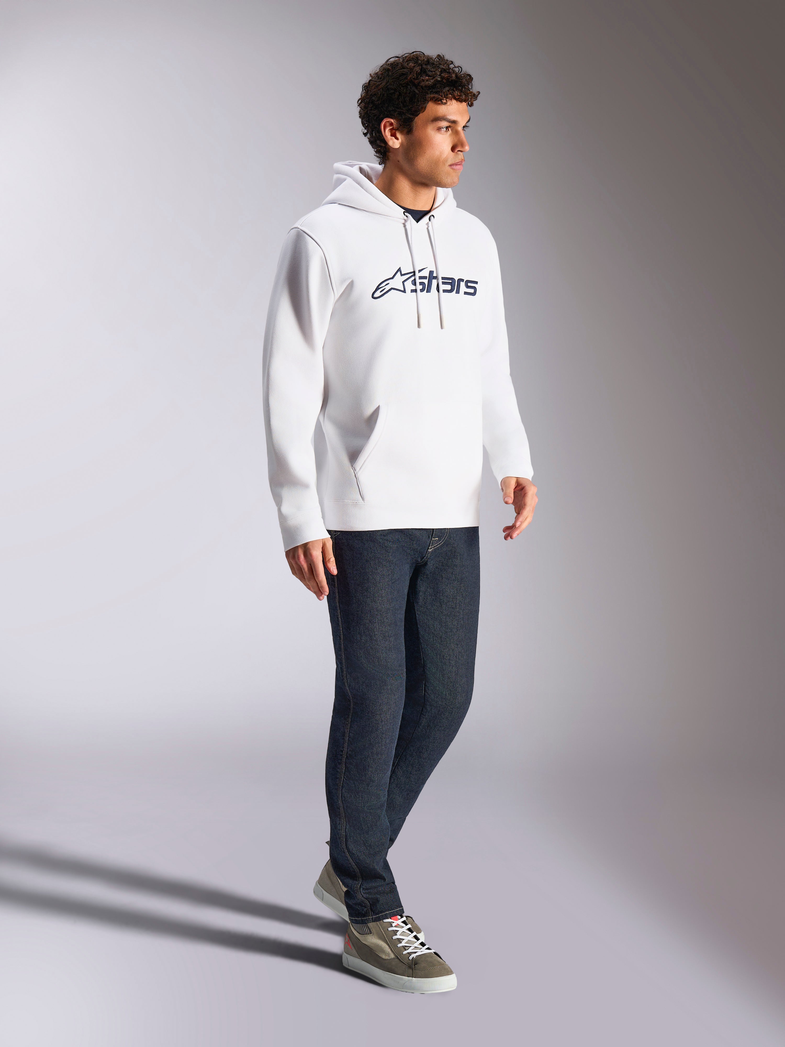 Alpinestars Blaze V3 Hoodie, branco e azul-marinho, streetwear casual usado por um modelo com calças de ganga azul-escuro e sapatilhas, com capuz de cordão e impressão marcante do logótipo na frente, design moderno inspirado no desporto motorizado