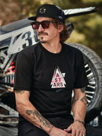 Boné Trucker Alpinestars Gambit em preto com remendo de logótipo circular, t-shirt Alpinestars Soloist SS em preto, imagem de estilo de vida de um homem com óculos de sol e tatuagens sentado em frente a uma mota de motocross.