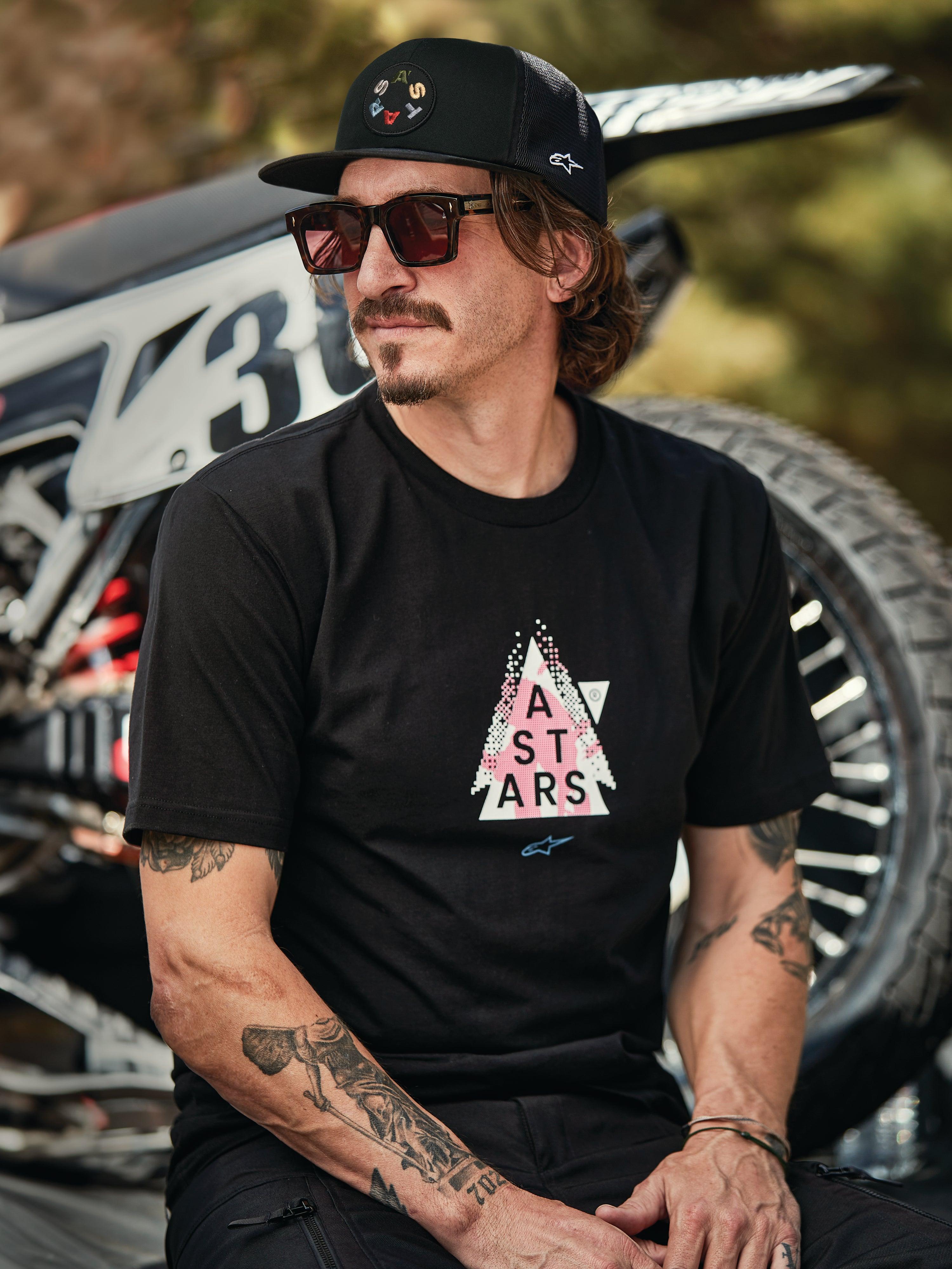 Boné Trucker Alpinestars Gambit em preto com remendo de logótipo circular, t-shirt Alpinestars Soloist SS em preto, imagem de estilo de vida de um homem com óculos de sol e tatuagens sentado em frente a uma mota de motocross.