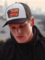 Boné Alpinestars Hardware Trucker em Military White, usado por um homem com um hoodie preto, painel frontal branco com aba curva e malha traseira pretas, remendo bordado bege com marca Alpinestars Motocross a vermelho, estilo de vida casual motorsport streetwear