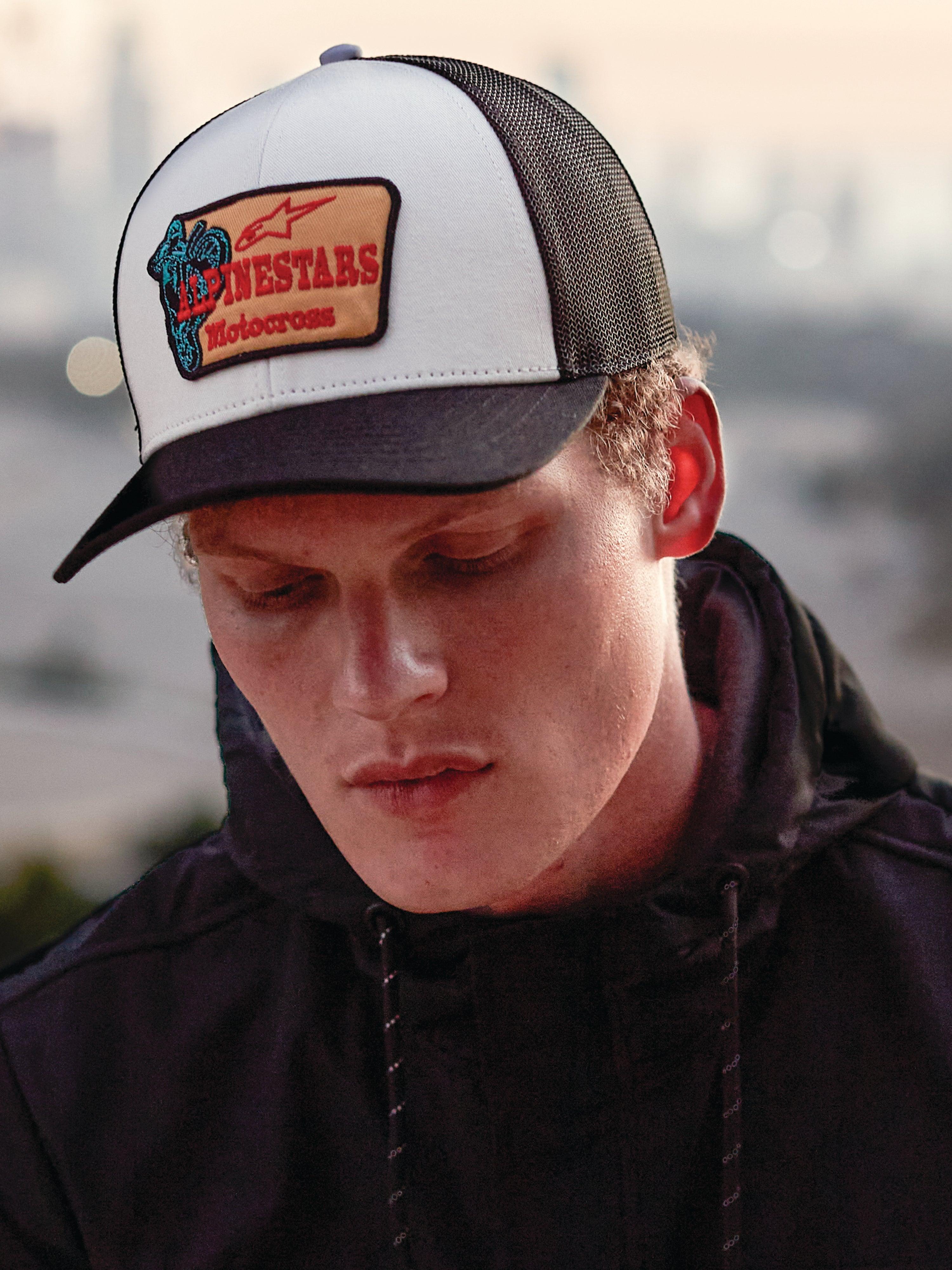 Boné Alpinestars Hardware Trucker em Military White, usado por um homem com um hoodie preto, painel frontal branco com aba curva e malha traseira pretas, remendo bordado bege com marca Alpinestars Motocross a vermelho, estilo de vida casual motorsport streetwear