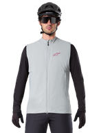 Colete Térmico Alpinestars A-Dura, colete de ciclismo, cinzento titanium, usado com uma camisola de manga comprida preta e capacete de ciclismo preto, logótipo da estrela Alpinestars vermelho no peito, leve e isolado para mountain biking.