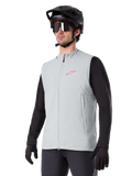 Colete Térmico Alpinestars A-Dura, cinzento Titanium, colete térmico de ciclismo, usado sobre uma camisola preta de manga comprida com capacete de BTT e luvas pretas, com logótipo rosa no peito e bolsos laterais verticais com fecho