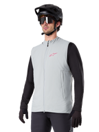 Colete Térmico Alpinestars A-Dura, cinzento Titanium, colete térmico de ciclismo, usado sobre uma camisola preta de manga comprida com capacete de BTT e luvas pretas, com logótipo rosa no peito e bolsos laterais verticais com fecho