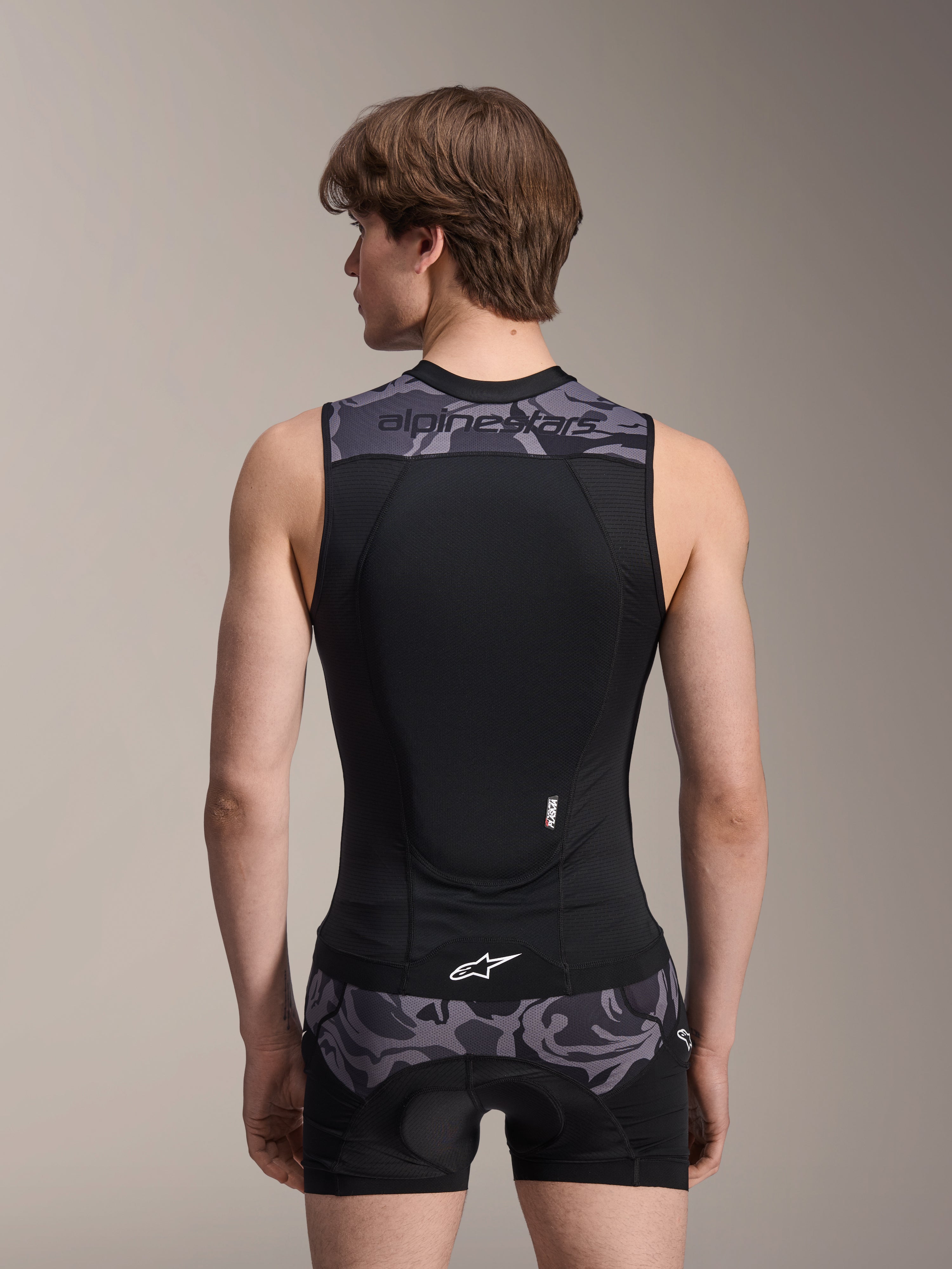 Colete de Proteção Alpinestars A-Motion Plasma, Black Camo, vista traseira num modelo masculino, com construção em malha respirável, protetor de costas Nucleon Plasma integrado, detalhes de camuflagem cinzenta nos ombros e na cintura, e logótipo Alpinestars.