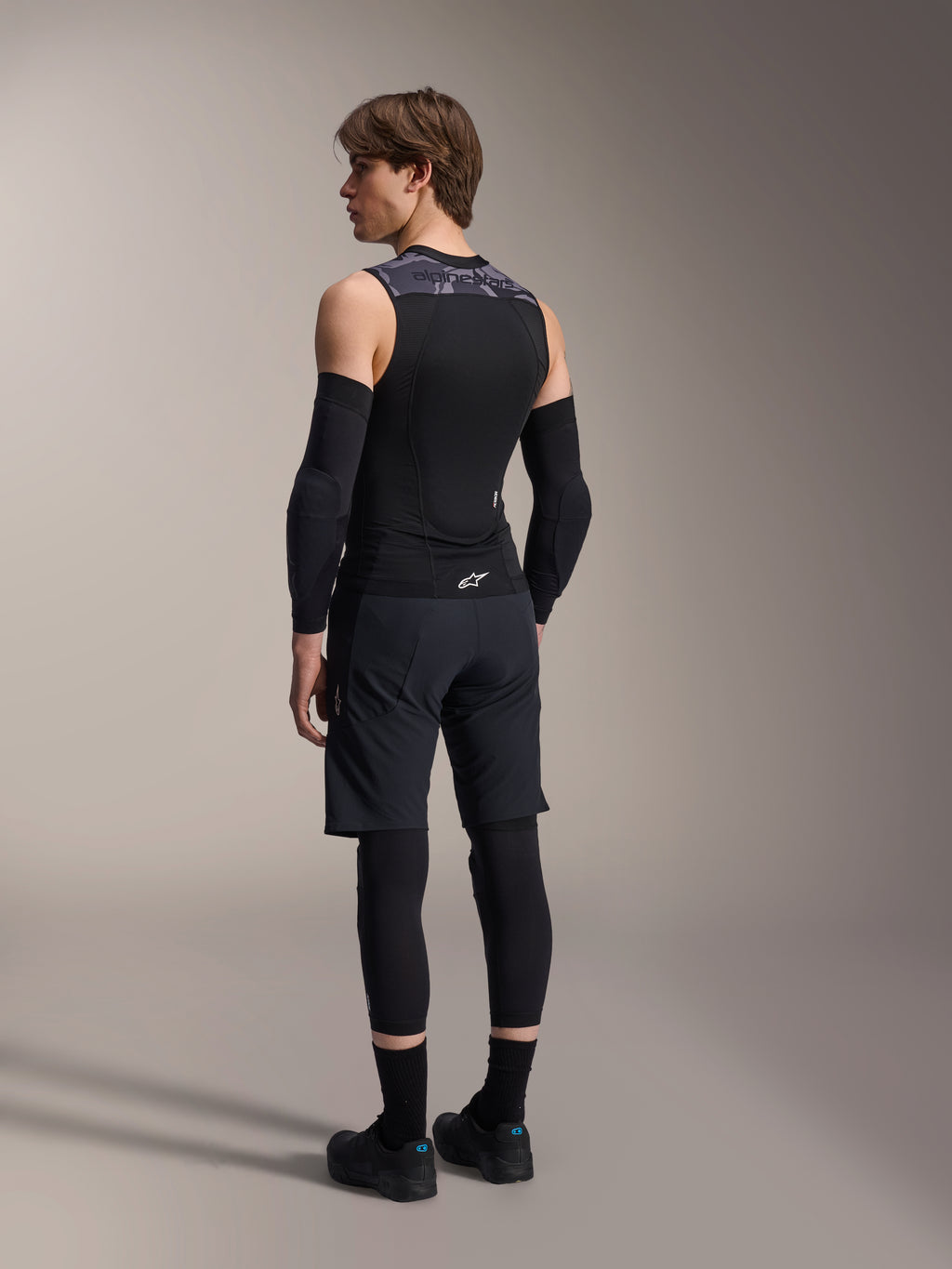 Modelo masculino visto de costas a usar o Colete de Proteção Alpinestars A-Motion Plasma, Black Camo, design em malha sem mangas com protetor de costas integrado, padrão camuflado cinzento nos ombros, usado com protetores de cotovelo pretos e calções de BTT, equipamento de ciclismo profissional.