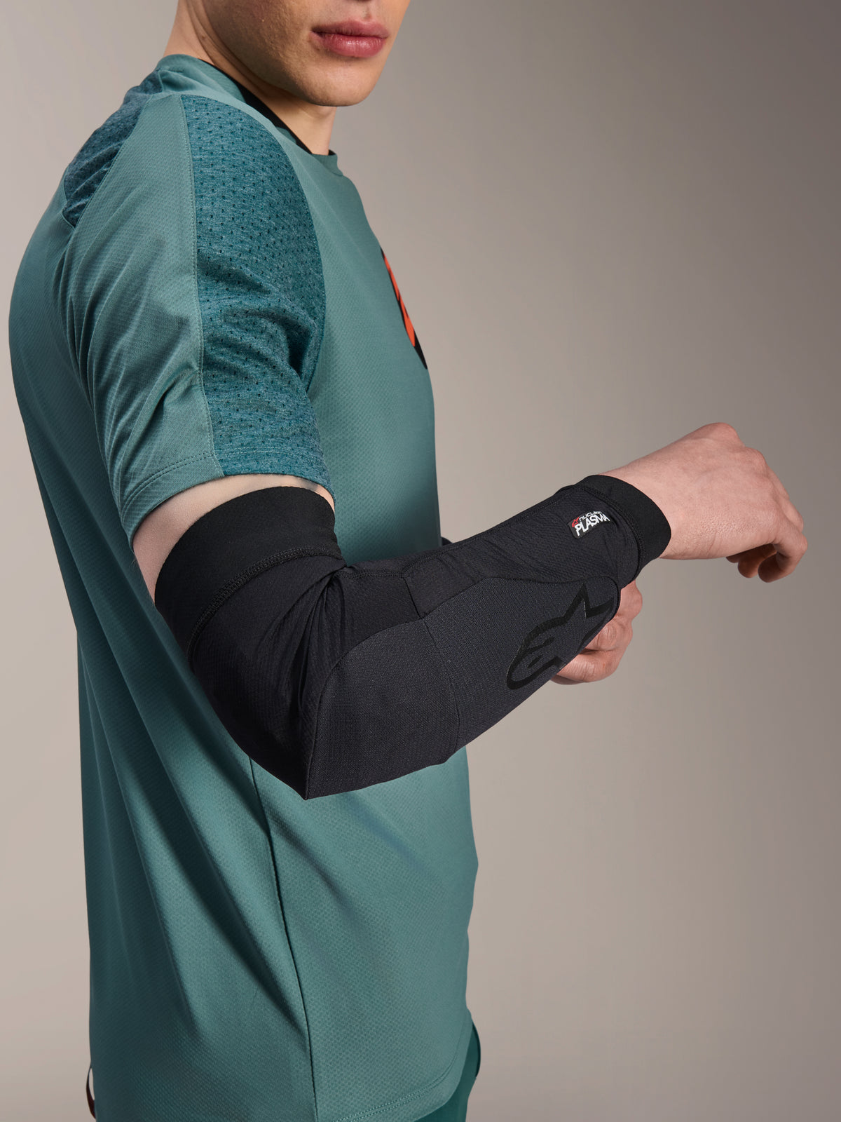 Cotoveleira Alpinestars A-Motion Plasma, protetor de cotovelo para ciclismo, preta, usada por um ciclista com uma camisola verde-azulado, com o logótipo Alpinestars e a marca Nucleon Plasma, design elegante e respirável para proteção de BTT