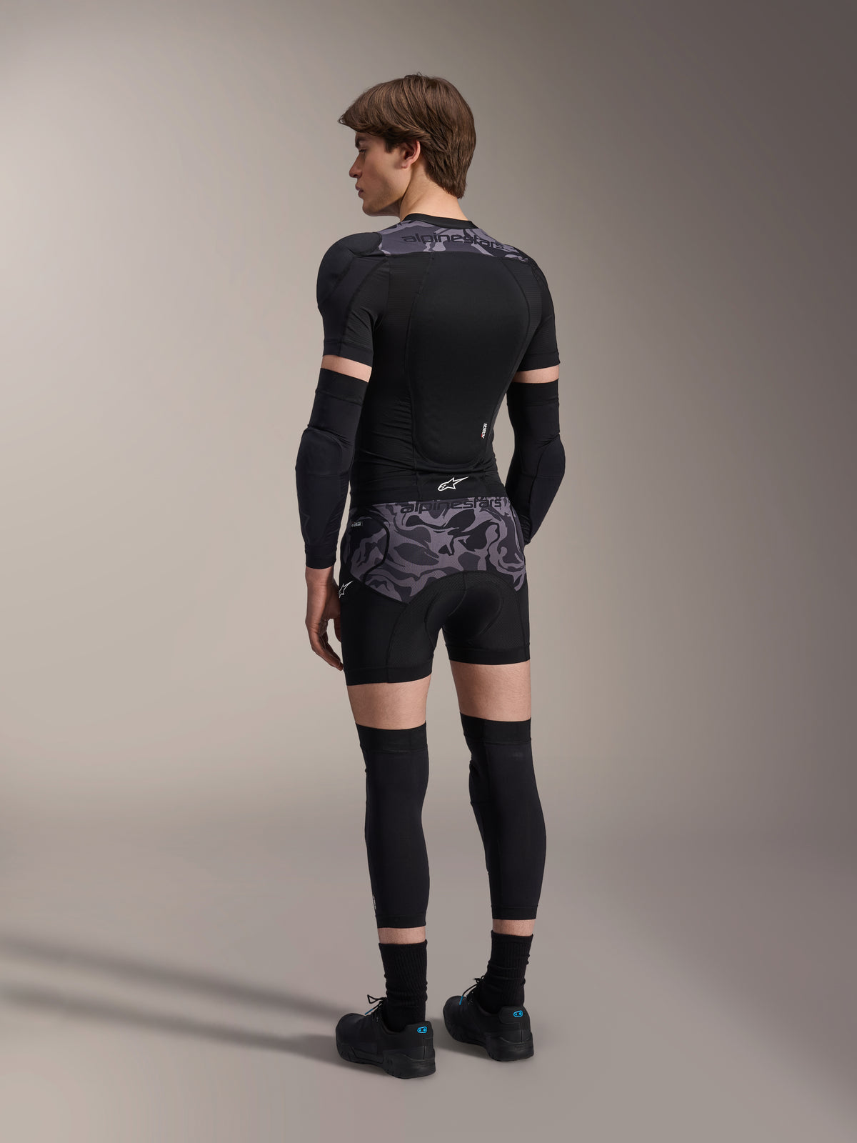 Modelo a usar os Boxers de Proteção Alpinestars A-Motion Plasma em Black Camo e o casaco de proteção A-Motion Plasma a condizer, vista traseira mostrando o padrão de camuflagem cinzento, cotoveleiras, joelheiras e sapatos de ciclismo, conjunto completo de equipamento de proteção para mountain bike