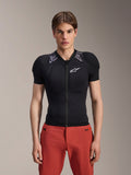 Modelo masculino a usar Alpinestars A-Motion Plasma Protection Jacket - Manga Curta, preto com acolchoamento de ombros em camuflado cinzento, logótipo Alpinestars branco no peito, combinado com calças vermelhas, armadura de proteção para BTT de corte justo com fecho de correr frontal