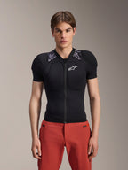 Modelo masculino a usar Alpinestars A-Motion Plasma Protection Jacket - Manga Curta, preto com acolchoamento de ombros em camuflado cinzento, logótipo Alpinestars branco no peito, combinado com calças vermelhas, armadura de proteção para BTT de corte justo com fecho de correr frontal