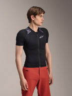 Modelo masculino a usar o Casaco de Proteção Alpinestars A-Motion Plasma de Manga Curta, em Black Camo com painéis de camuflagem cinzenta nos ombros, com protetores de peito e ombros integrados, logótipo de estrela branco no peito, tecido de malha respirável, combinado com calças vermelhas