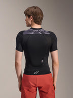 Casaco de Proteção Alpinestars A-Motion Plasma - Manga Curta, preto com detalhes de camuflagem cinzenta na parte superior das costas, vista traseira mostrando o protetor de costas integrado e o logótipo Astars branco na bainha, usado com calções vermelhos, equipamento técnico de proteção para BTT