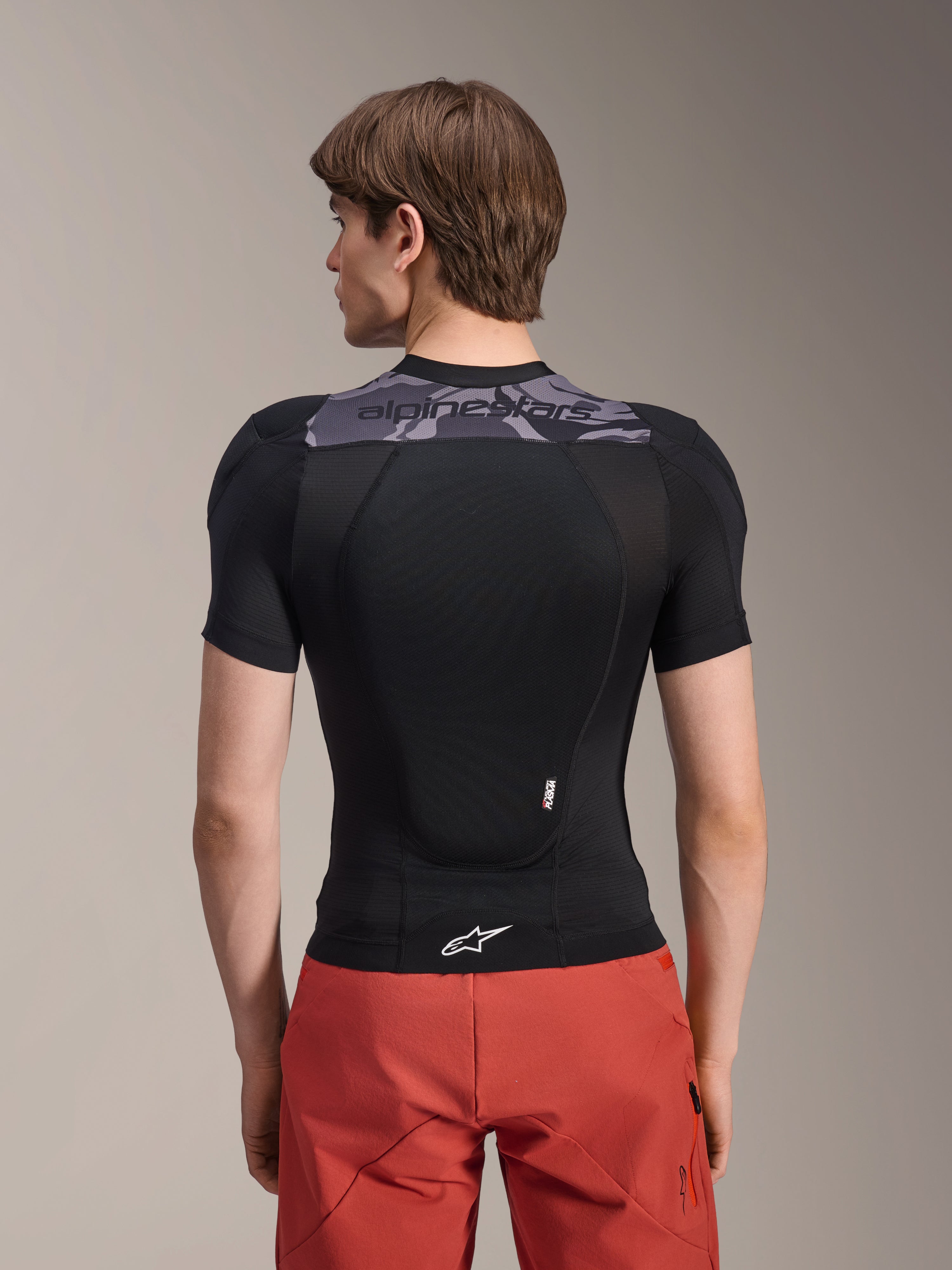 Casaco de Proteção Alpinestars A-Motion Plasma - Manga Curta, preto com detalhes de camuflagem cinzenta na parte superior das costas, vista traseira mostrando o protetor de costas integrado e o logótipo Astars branco na bainha, usado com calções vermelhos, equipamento técnico de proteção para BTT