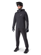 Male mountain biker wearing Alpinestars A-Dura Waterproof Pants in black, paired with a matching black hooded waterproof jacket, black MTB helmet with visor, dark cycling glasses, and black gloves, full professional gear set for all-weather trail riding