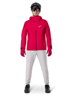 Calças Impermeáveis Alpinestars A-Dura, calças de BTT, cinzento Titanium, usadas com um casaco vermelho vivo com capuz, luvas vermelhas e um capacete de BTT preto, equipamento de ciclismo profissional impermeável para condições de trilhos molhados