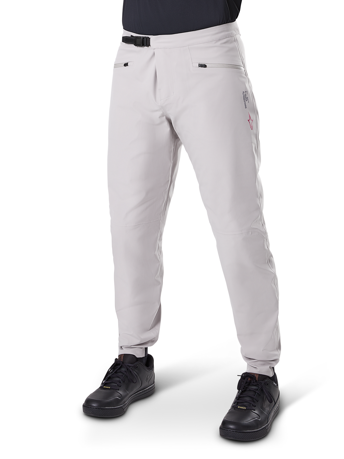Calças Impermeáveis Alpinestars A-Dura, calças para BTT, cinza Titanium, com fecho de catraca preto na cintura e bolsos de mão com fecho de correr, pernas afuniladas com punhos elásticos, usadas com sapatos pretos, construção impermeável 3L durável para ciclismo fora de estrada