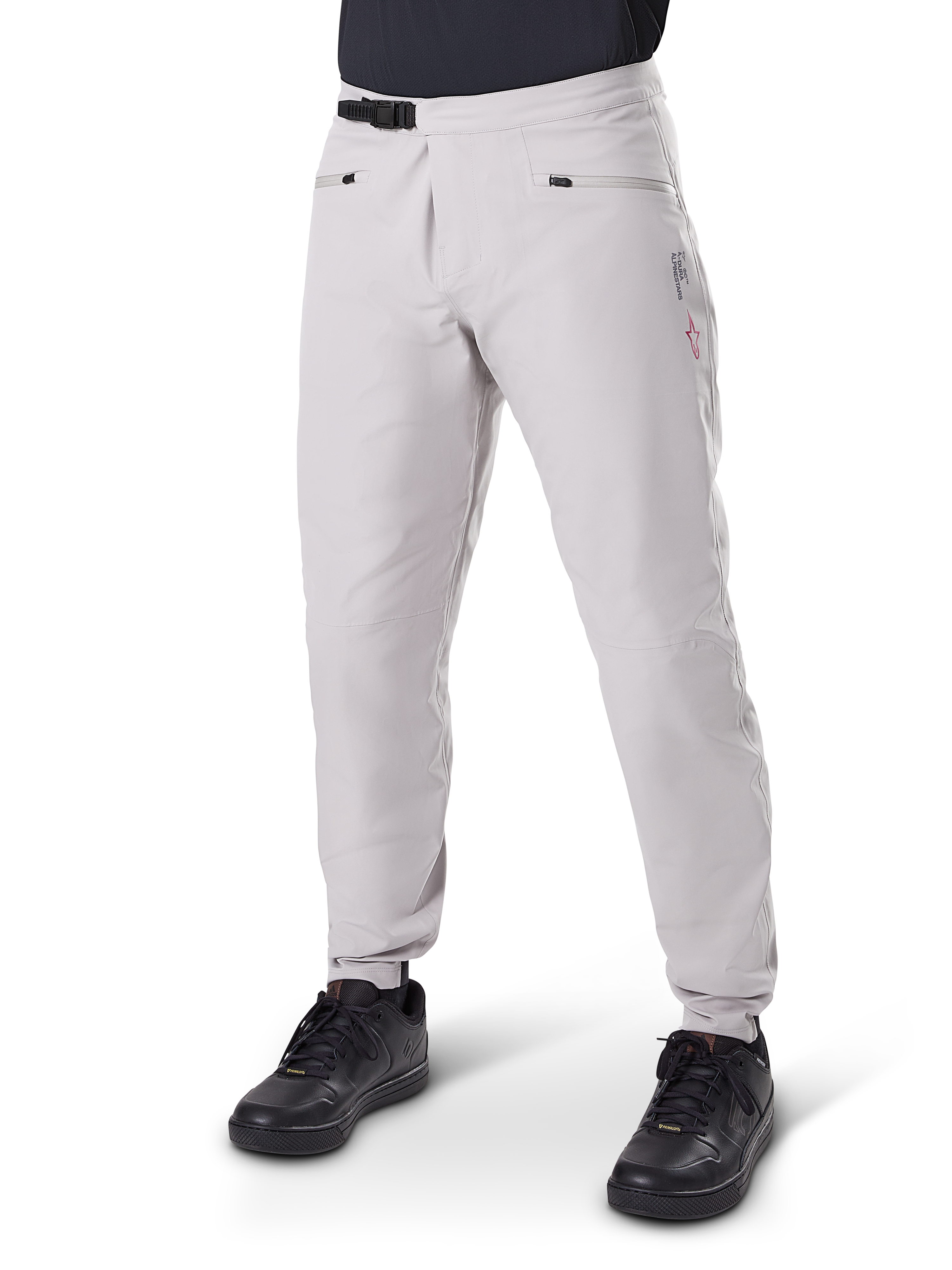 Calças Impermeáveis Alpinestars A-Dura, calças para BTT, cinza Titanium, com fecho de catraca preto na cintura e bolsos de mão com fecho de correr, pernas afuniladas com punhos elásticos, usadas com sapatos pretos, construção impermeável 3L durável para ciclismo fora de estrada