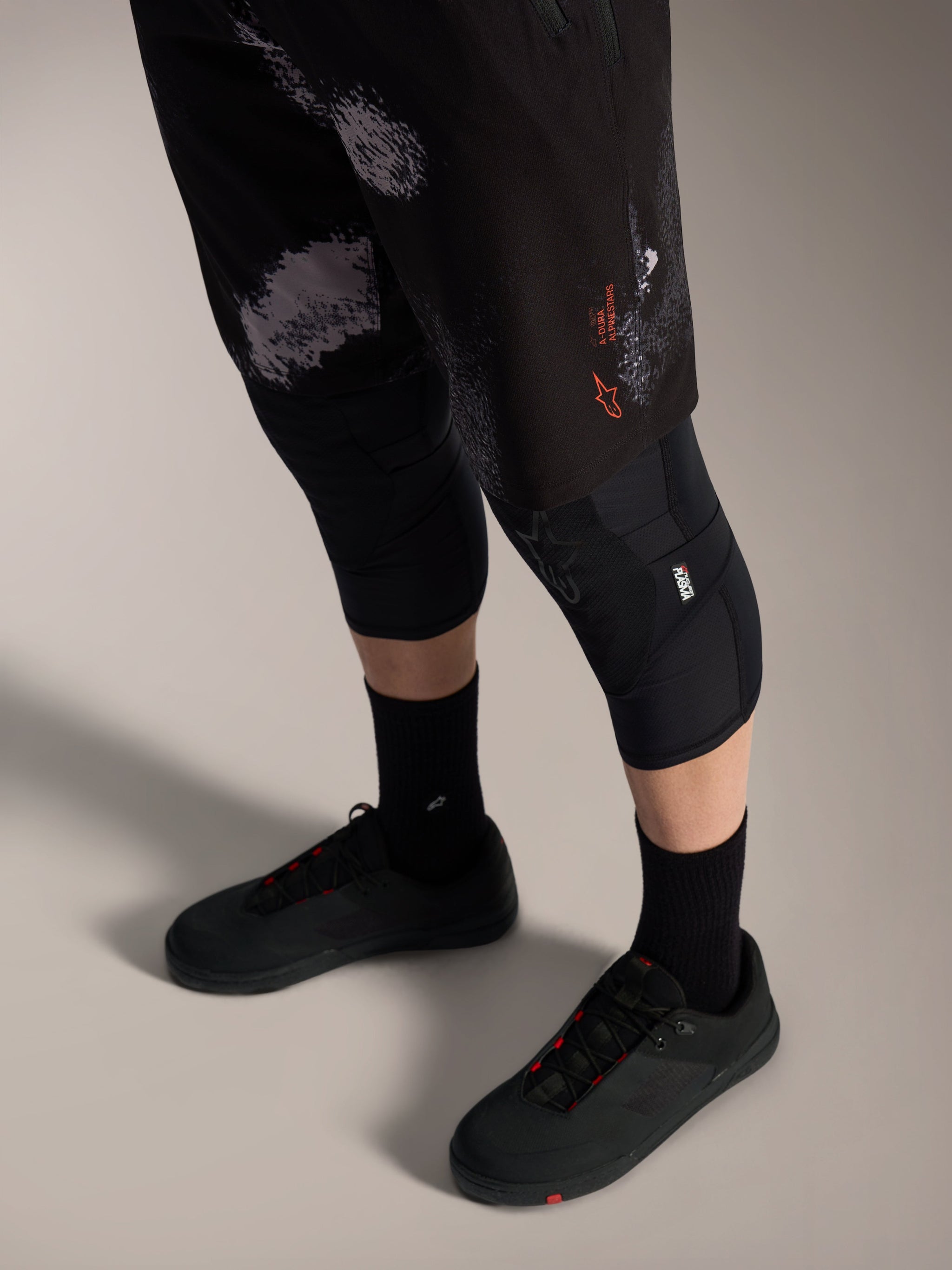 Alpinestars A-Dura Ride Shorts in Black Camo, durable MTB gear with orange logo, worn with knee guards and cycling shoes.