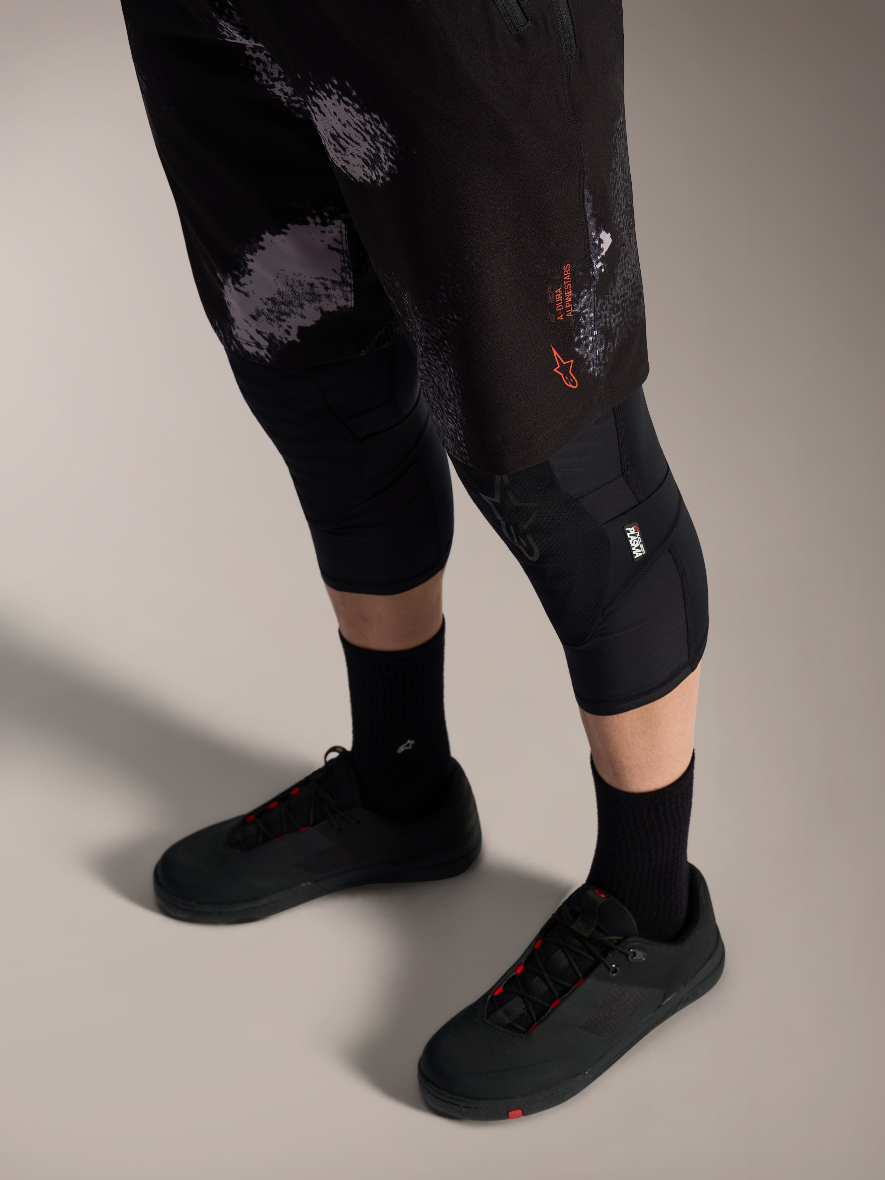 Calções Alpinestars A-Dura Ride Black Camo, equipamento de BTT com logótipo laranja, joelheiras e sapatos de ciclismo.