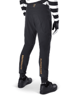 Alpinestars A-Aria Elite Pants, calças de BTT pretas, vista traseira, com logótipo Alpinestars vertical em dourado na parte de trás das pernas, usadas com camisola às riscas pretas e brancas e sapatos pretos, vestuário de performance para ciclismo de montanha