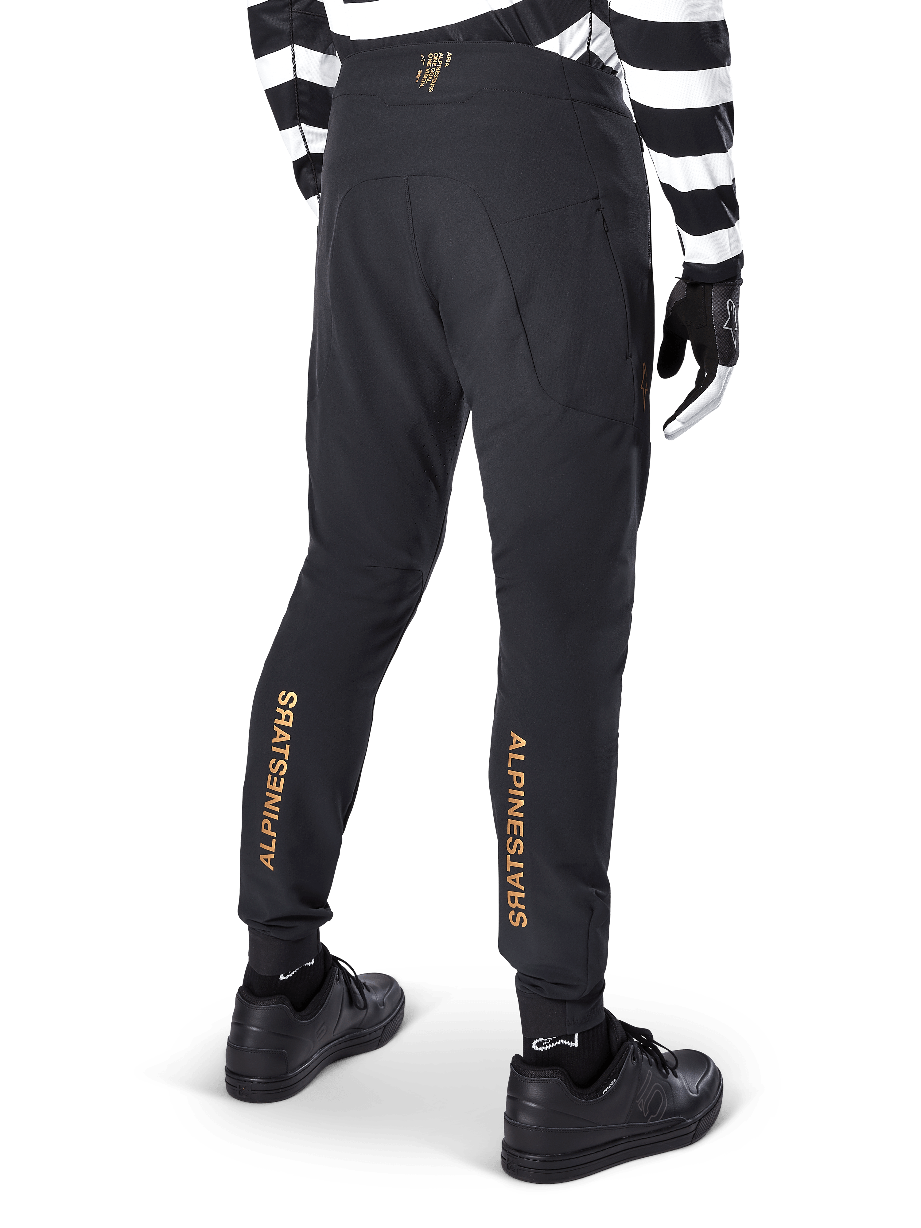 Alpinestars A-Aria Elite Pants, calças de BTT pretas, vista traseira, com logótipo Alpinestars vertical em dourado na parte de trás das pernas, usadas com camisola às riscas pretas e brancas e sapatos pretos, vestuário de performance para ciclismo de montanha