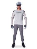 Alpinestars A-Aria Elite Pants, Dark Gray Mountain Bike Pants, worn by a rider in a light gray long-sleeve jersey, light blue helmet, and black shoes, featuring visible laser perforations on the inner legs and a slim performance fit.