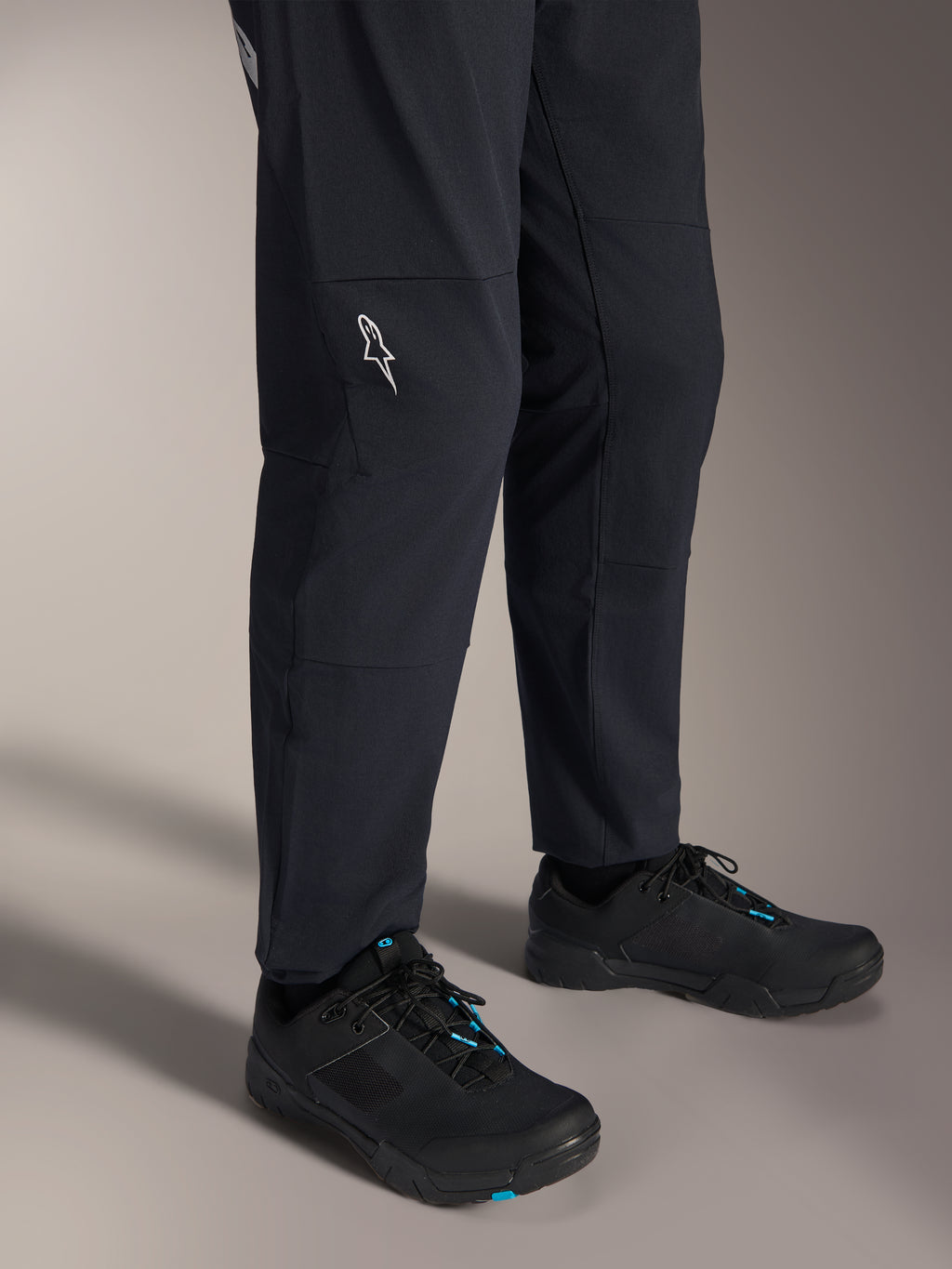 Alpinestars A-Dura Elite Pants, calças de mountain bike pretas, vista da parte inferior das pernas com logótipo Alpinestars branco no joelho, usadas com sapatos de ciclismo pretos e azuis, construção durável para a prática de MTB