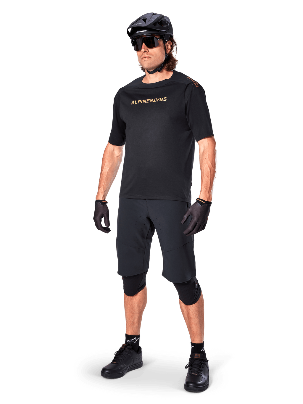 Ciclista a usar Calções Alpinestars A-Aria Elite, calções de BTT pretos, combinados com uma camisola preta com logótipo dourado, capacete preto, óculos de sol e joelheiras, equipamento de performance completo para ciclismo fora de estrada
