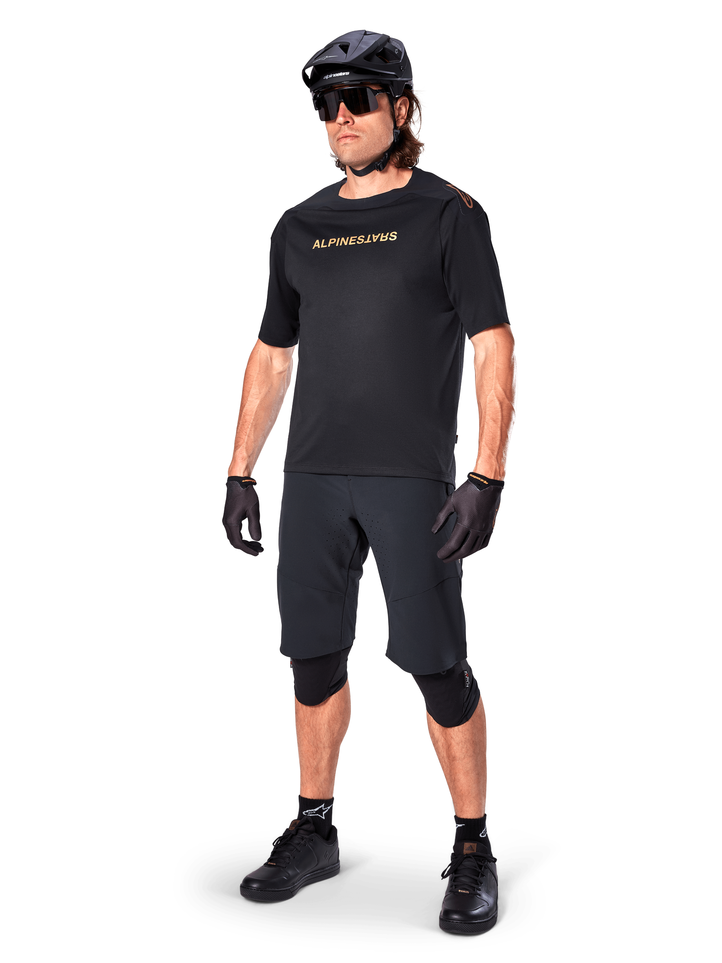 Ciclista a usar Calções Alpinestars A-Aria Elite, calções de BTT pretos, combinados com uma camisola preta com logótipo dourado, capacete preto, óculos de sol e joelheiras, equipamento de performance completo para ciclismo fora de estrada