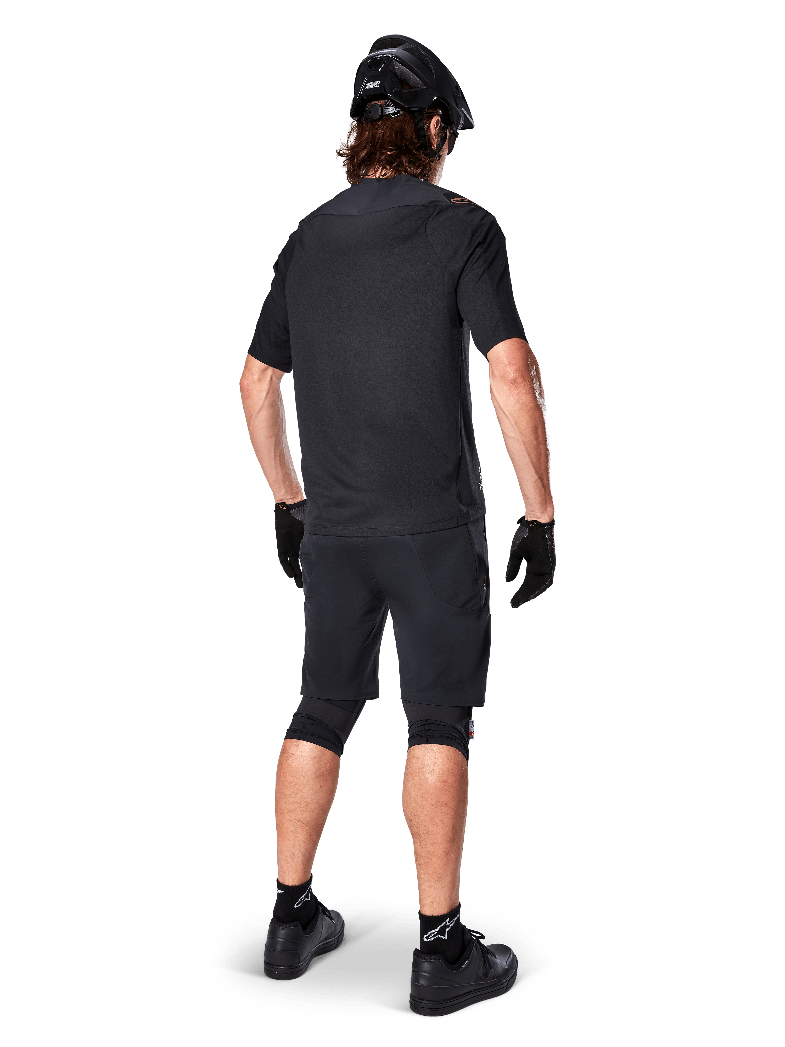 Vista traseira de um ciclista de montanha a usar calções Alpinestars A-Aria Elite em preto, combinados com uma camisola de manga curta preta a condizer, capacete preto, luvas e sapatos de BTT, mostrando um equipamento de performance completo para ciclismo fora de estrada.