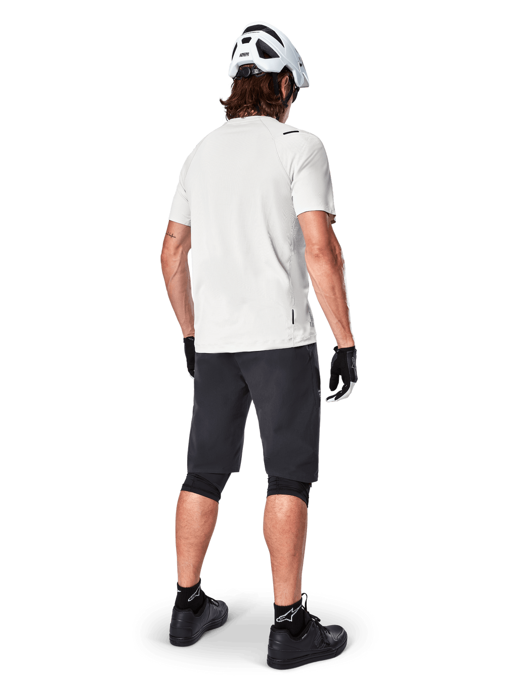 Ciclista de montanha visto de trás a usar calções Alpinestars A-Aria pretos, camisola de performance branca de manga curta, capacete branco, luvas pretas e sapatos de BTT pretos, equipamento de ciclismo para trilhos