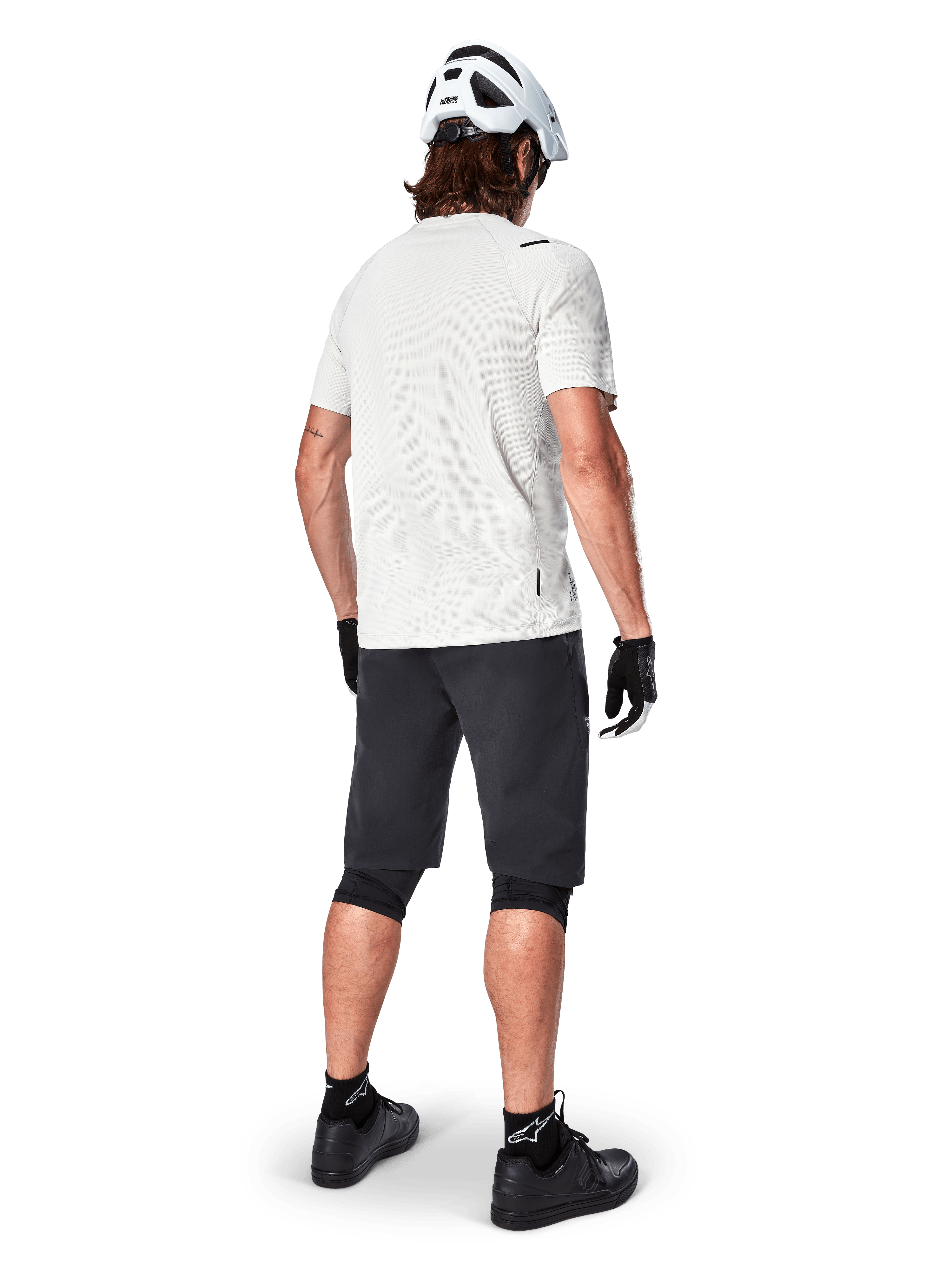Ciclista de montanha visto de trás a usar calções Alpinestars A-Aria pretos, camisola de performance branca de manga curta, capacete branco, luvas pretas e sapatos de BTT pretos, equipamento de ciclismo para trilhos