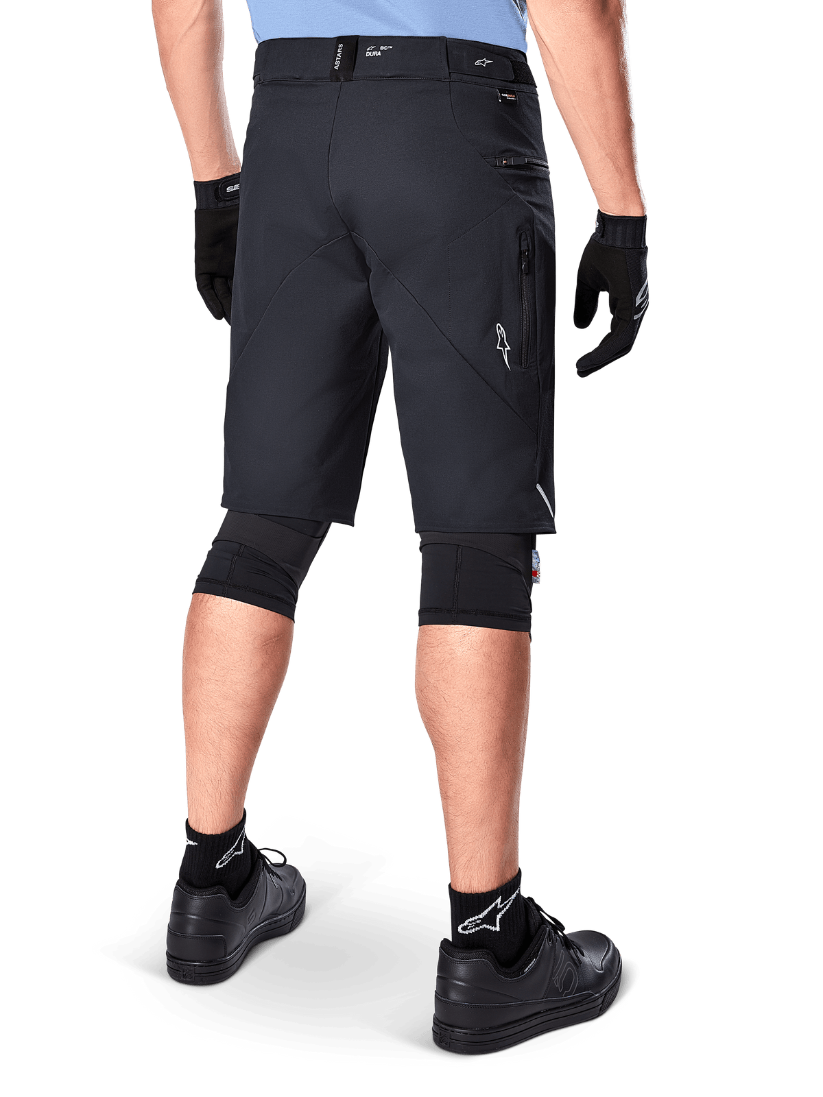 Calções Alpinestars A-Dura Elite, pretos, vista traseira de um ciclista com equipamento de mountain bike, incluindo joelheiras pretas, luvas e meias com logótipos brancos, com bolsos laterais com fecho e detalhes refletores em tecido Cordura resistente.