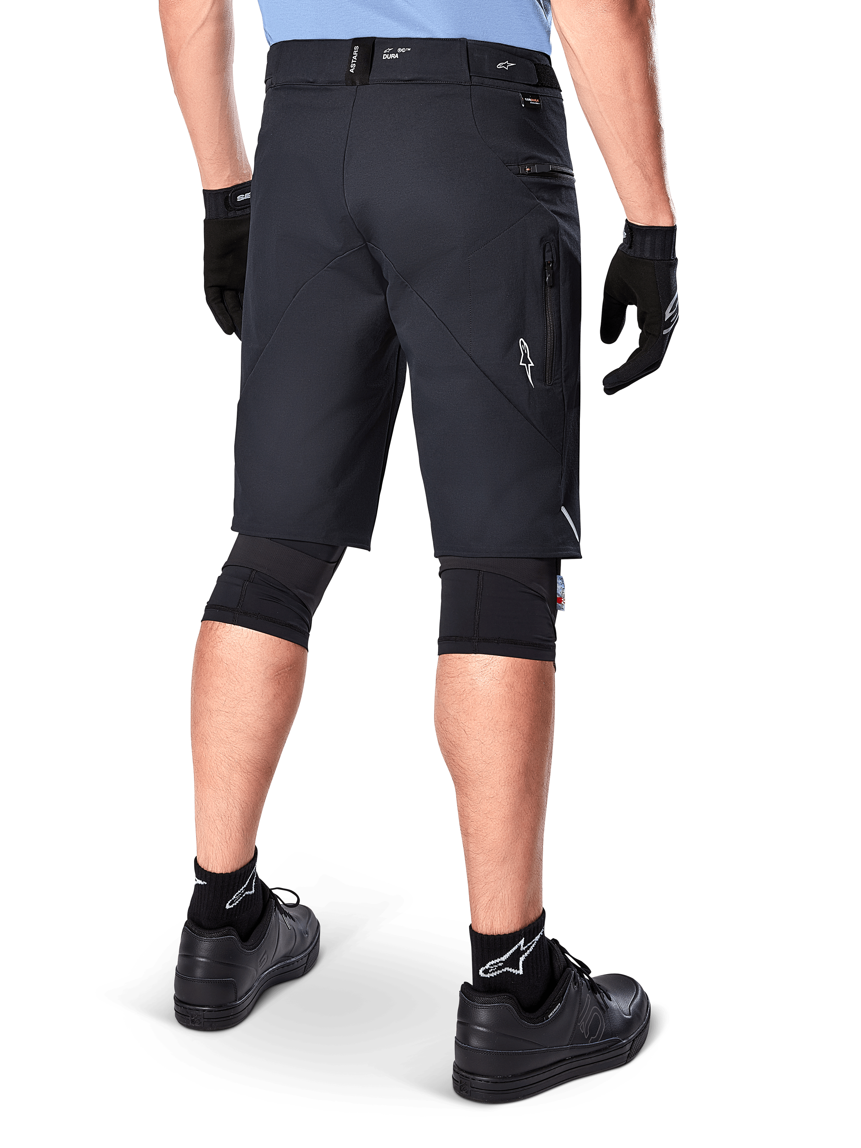 Calções Alpinestars A-Dura Elite, pretos, vista traseira de um ciclista com equipamento de mountain bike, incluindo joelheiras pretas, luvas e meias com logótipos brancos, com bolsos laterais com fecho e detalhes refletores em tecido Cordura resistente.