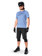 Ciclista masculino com calções Alpinestars A-Dura Elite em preto, camisola de manga curta azul claro com logótipo no peito, capacete preto, óculos de sol e joelheiras de proteção, equipamento completo de mountain bike para trilhos fora de estrada