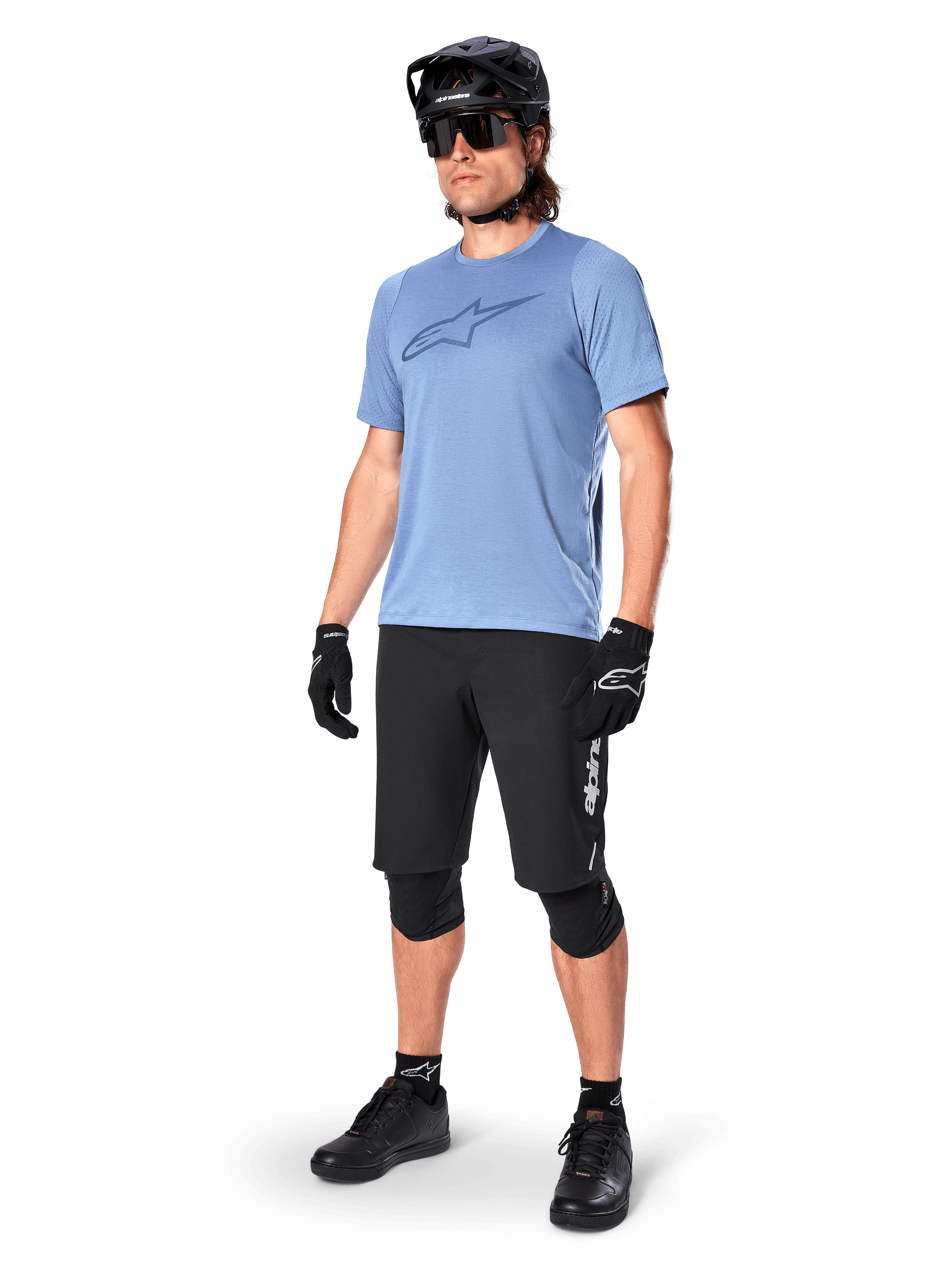 Ciclista masculino com calções Alpinestars A-Dura Elite em preto, camisola de manga curta azul claro com logótipo no peito, capacete preto, óculos de sol e joelheiras de proteção, equipamento completo de mountain bike para trilhos fora de estrada
