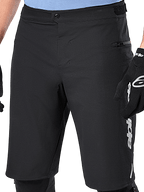 Calções Alpinestars A-Dura Elite, calções de BTT pretos, vista de pormenor com fecho de pressão frontal e bolso lateral com fecho de correr, usados por um ciclista com luvas de ciclismo pretas e joelheiras, equipamento de alta performance para BTT