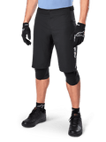 Alpinestars A-Dura Elite Shorts, calções de BTT pretos, usados com camisola azul clara e proteção de joelhos preta, com fecho de pressão seguro e bolso com fecho de correr, combinados com sapatos de ciclismo e luvas pretas para um visual completo de todo-o-terreno.