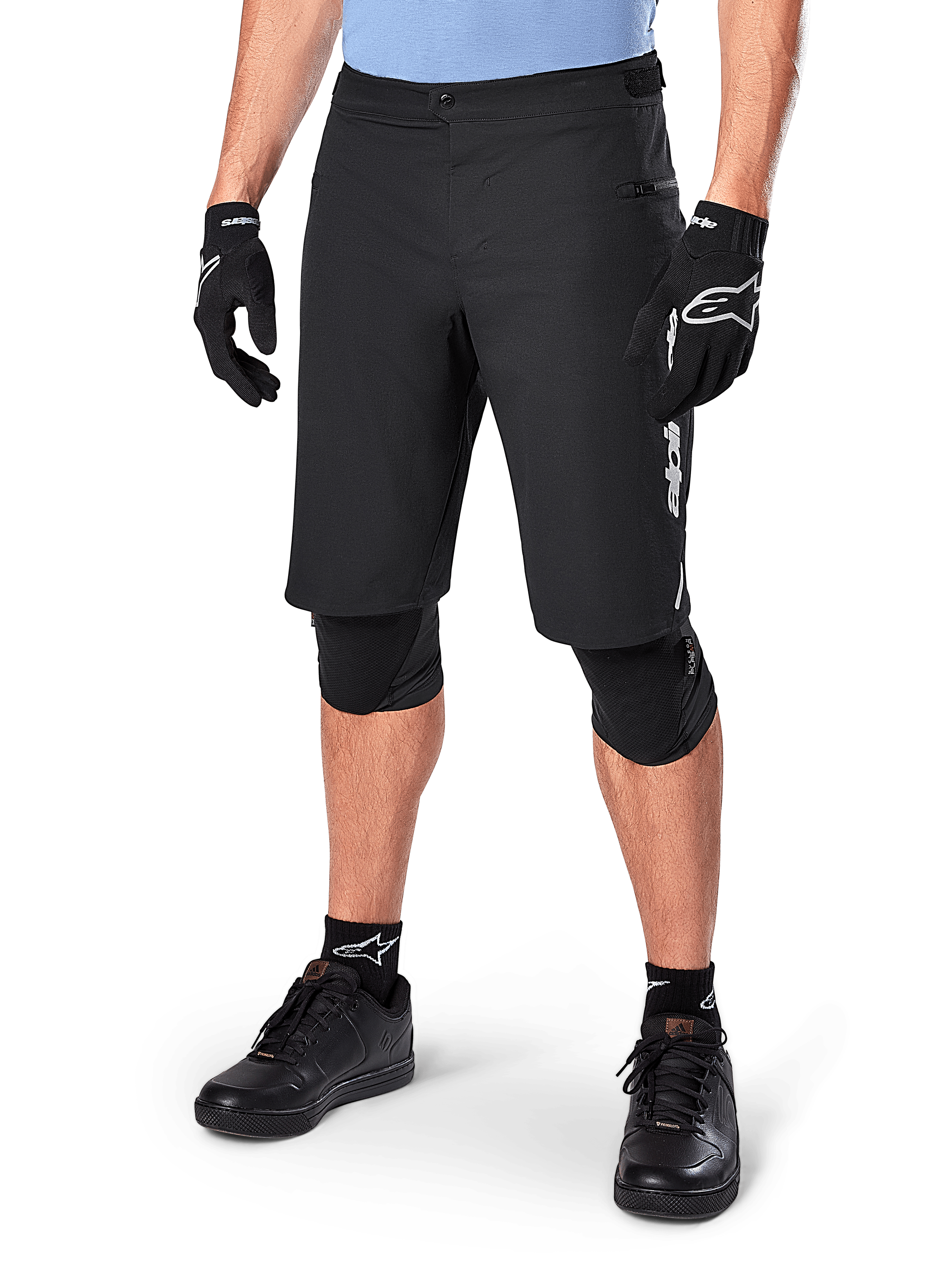 Alpinestars A-Dura Elite Shorts, calções de BTT pretos, usados com camisola azul clara e proteção de joelhos preta, com fecho de pressão seguro e bolso com fecho de correr, combinados com sapatos de ciclismo e luvas pretas para um visual completo de todo-o-terreno.