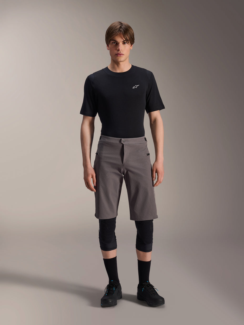 Alpinestars A-Dura Elite Shorts, calções de BTT cinzento escuro, usados com t-shirt preta de gola redonda e joelheiras pretas, vestuário de ciclismo de performance pelo joelho com fecho frontal de mola e tecido Cordura durável