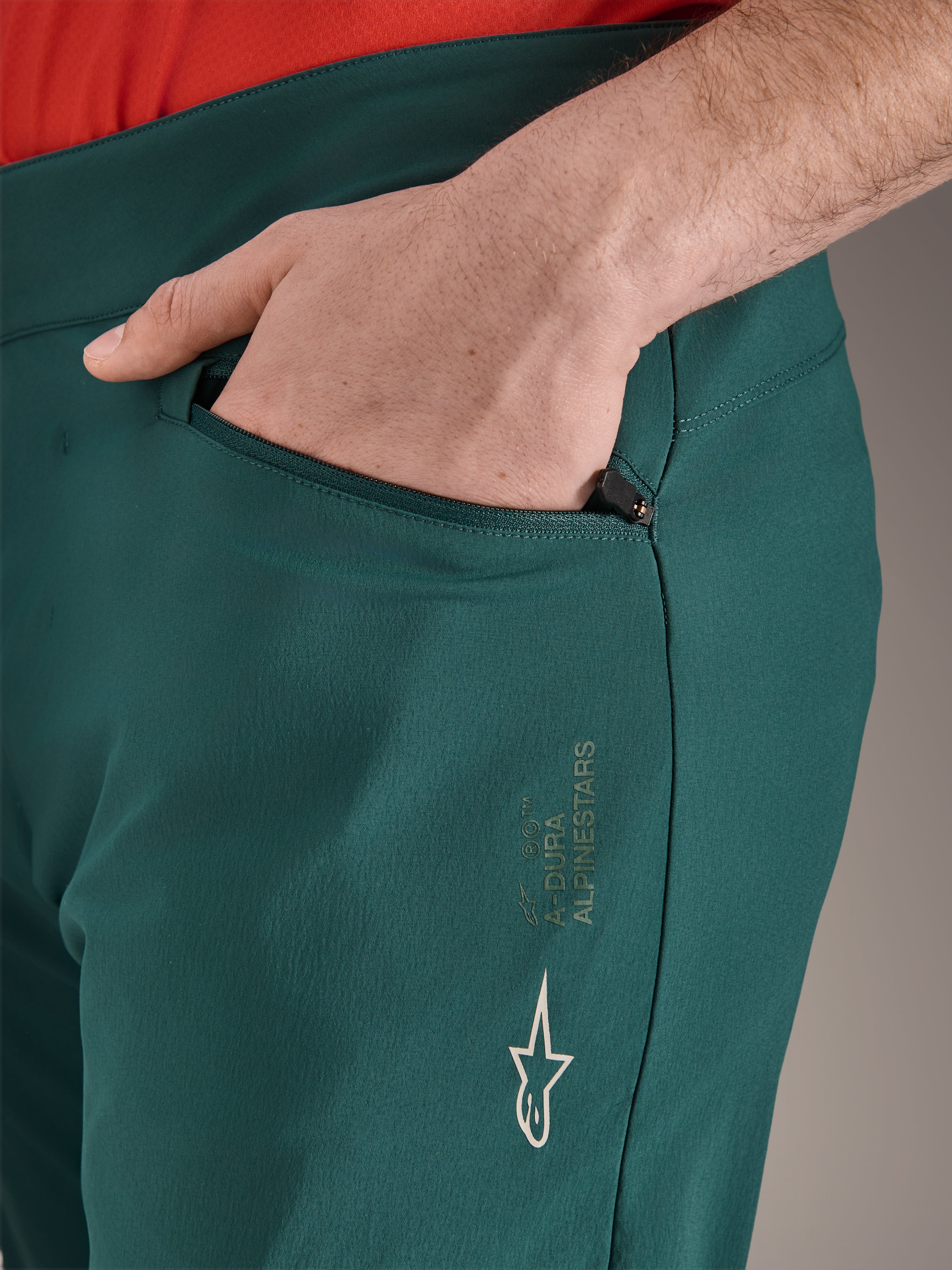 Calças Alpinestars A-Dura em verde Dark Pine, grande plano do bolso com fecho de correr e mão, com o logótipo e a marca Alpinestars em branco na coxa, calças de ciclismo MTB duráveis