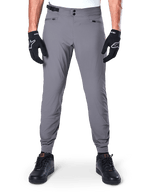 Calças Alpinestars A-Dura, calças de BTT cinzento-escuro, vista frontal, usadas com luvas pretas e ténis pretos, com fecho de cintura de pressão duplo, bolsos com fecho de correr e punhos elásticos, vestuário de ciclismo durável para trail