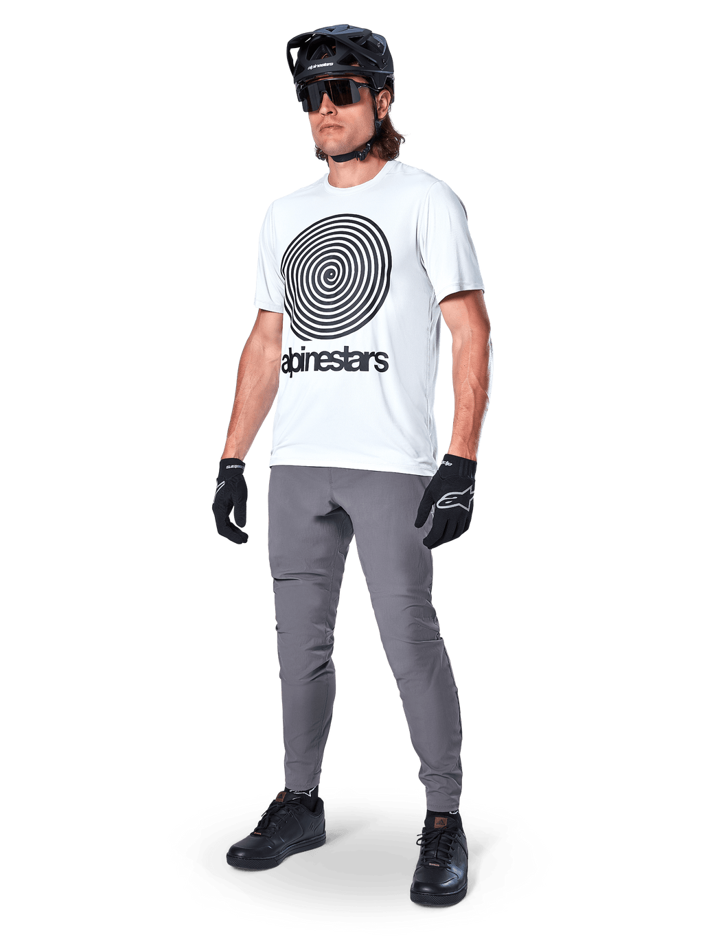Calças Alpinestars A-Dura, calças de MTB, cinza escuro, usadas com t-shirt branca com grafismo em espiral, capacete de MTB preto, óculos de sol e luvas pretas, tecido durável com elasticidade em 2 direções para ciclismo fora de estrada