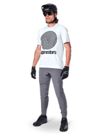 Calças Alpinestars A-Dura, calças de MTB, cinza escuro, usadas com t-shirt branca com grafismo em espiral, capacete de MTB preto, óculos de sol e luvas pretas, tecido durável com elasticidade em 2 direções para ciclismo fora de estrada
