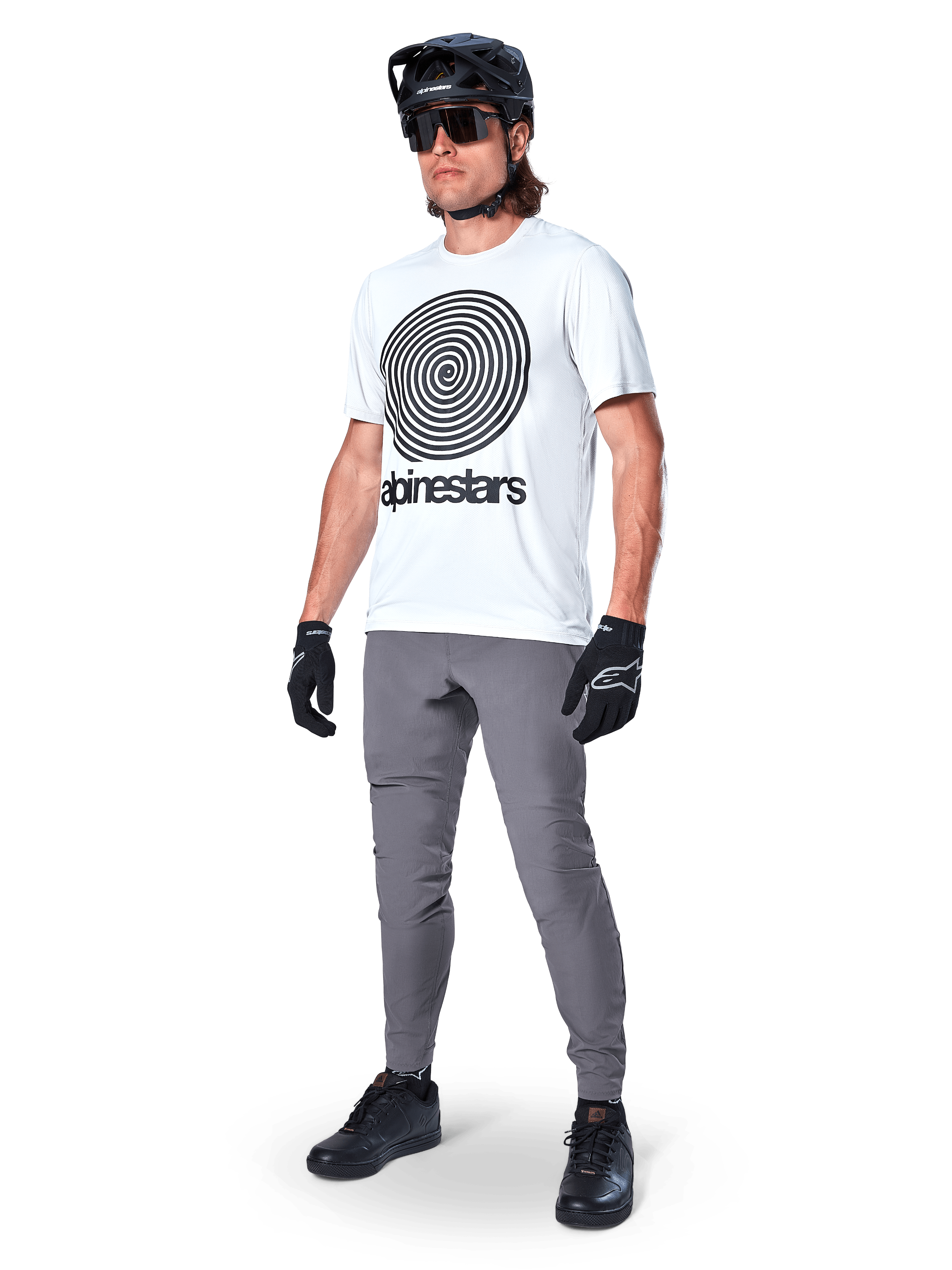 Calças Alpinestars A-Dura, calças de MTB, cinza escuro, usadas com t-shirt branca com grafismo em espiral, capacete de MTB preto, óculos de sol e luvas pretas, tecido durável com elasticidade em 2 direções para ciclismo fora de estrada
