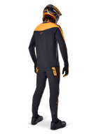 Alpinestars A-Supra Race Pants, MTB Racing Pants, Black with orange logos, viewed from the back on a rider wearing a matching black and orange jersey, helmet, and gloves, highlighting the MX-inspired wordmark on the back yoke and performance fit for technical trails.